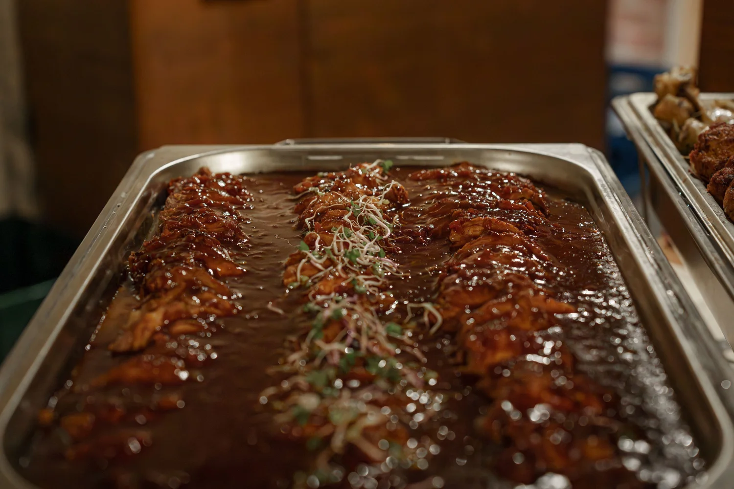 Königliche Schlossgastronomie – Dieses Bild zeigt ein Gericht aus glasiertem Fleisch, wahrscheinlich Schweinerippchen oder Hühnchen in einer dicken, dunklen Barbecue-Sauce, serviert in einer großen Catering-Chafer-Dish aus Edelstahl.